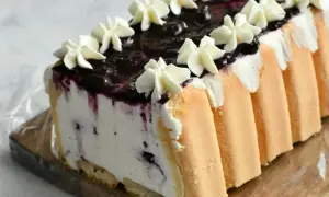 Pyszne ciasto śmietanowiec bez pieczenia z borówkami i bitą śmietaną, otulone biszkoptem.