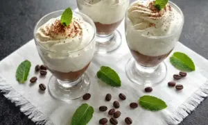 Trzy pucharki z deserem caffe latte, udekorowane bitą śmietaną, kakao i listkami mięty, otoczone ziarnami kawy.