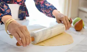 Ręce z biżuterią wałkują ciasto francuskie na papierze do pieczenia. W tle gruszki i jabłka.