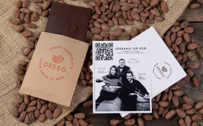 Czekolada DESEO, od ziarna do tabliczki (bean to bar), otoczona ziarnami kakaowca. Obok opakowanie i ulotka z QR kodem.