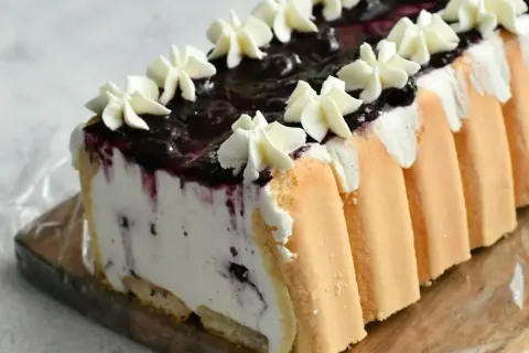 Pyszne ciasto śmietanowiec bez pieczenia z borówkami i bitą śmietaną, otulone biszkoptem.