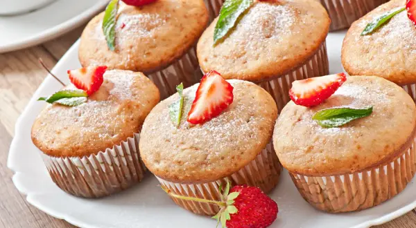 Słodkie muffinki z truskawkami i listkami mięty, oprószone cukrem pudrem, wyglądają jakby były polane polewą z serka mascarpone.