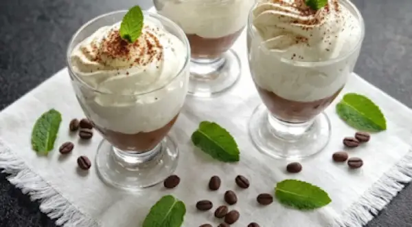 Trzy pucharki z deserem caffe latte, udekorowane bitą śmietaną, kakao i listkami mięty, otoczone ziarnami kawy.