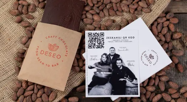 Czekolada DESEO, od ziarna do tabliczki (bean to bar), otoczona ziarnami kakaowca. Obok opakowanie i ulotka z QR kodem.
