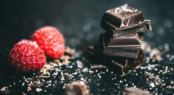 Kawałki ciemnej belgijskiej czekolady w tabliczkach, posypane solą, obok świeżych malin.