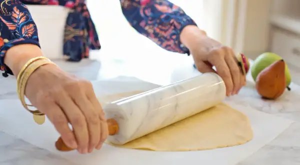 Ręce z biżuterią wałkują ciasto francuskie na papierze do pieczenia. W tle gruszki i jabłka.