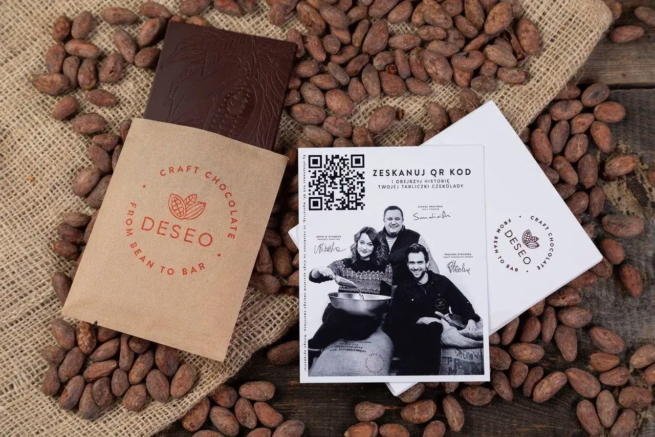 Czekolada DESEO, od ziarna do tabliczki (bean to bar), otoczona ziarnami kakaowca. Obok opakowanie i ulotka z QR kodem.