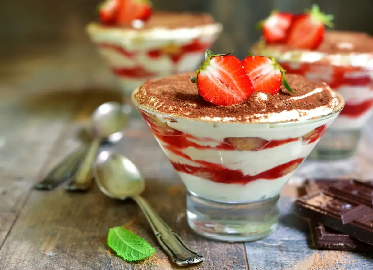 Pyszne tiramisu z truskawkami i polewą z serka mascarpone, posypane kakao. Obok leżą łyżeczki i kawałki czekolady.