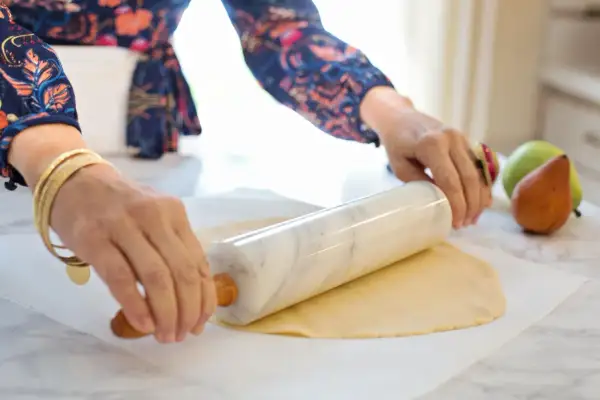 Ręce z biżuterią wałkują ciasto francuskie na papierze do pieczenia. W tle gruszki i jabłka.
